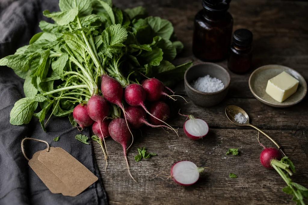 A bunch of red radishes with leafy tops, a few halved, beside flaky salt and butter on dark wood