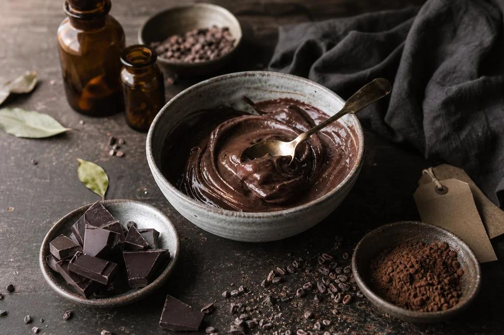 A stoneware bowl of dark chocolate pudding beside broken dark chocolate and scattered cacao nibs
