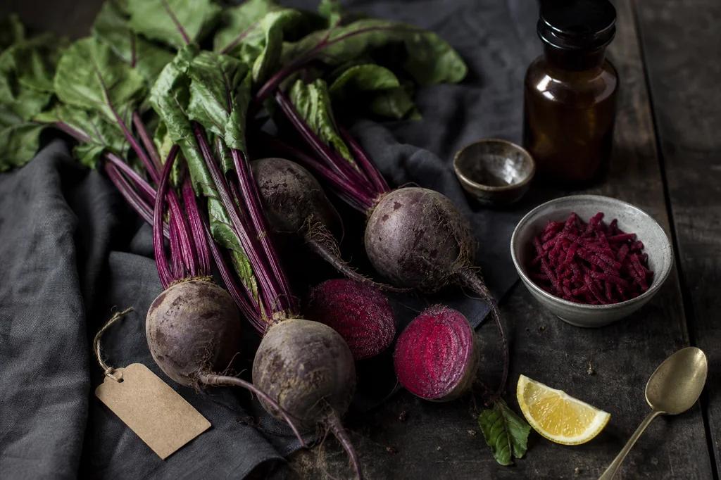 Whole beetroots with leafy stems, one cut open to show magenta rings, beside a bowl of grated beet and a lemon wedge